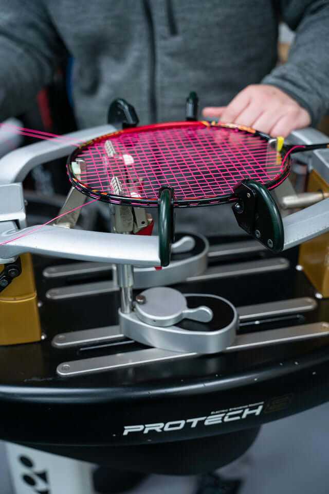 Racket Restringing Service with a black badminton racket fitted with bright pink strings on a stringing machine